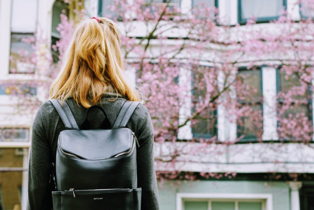 Person wearing school bag facing building and tree with pink leaves